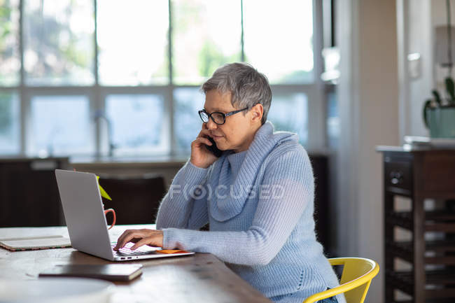 Вид сбоку на взрослую белую женщину с короткими седыми волосами в очках, сидящую за обеденным столом и делающую телефонный звонок на смартфоне с помощью ноутбука — стоковое фото