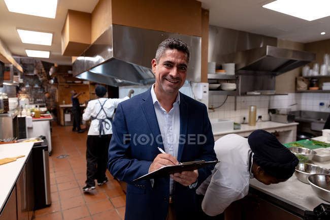 Retrato de cerca de un hombre de mediana edad gerente de restaurante caucásico escribiendo en un portapapeles en una cocina de restaurante ocupada, mientras que el personal de cocina trabaja en el fondo - foto de stock