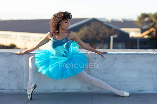 Vista frontal de una joven bailarina de ballet de raza mixta con un tutú azul sosteniendo una posición de ballet y mirando hacia la azotea de un edificio urbano - foto de stock