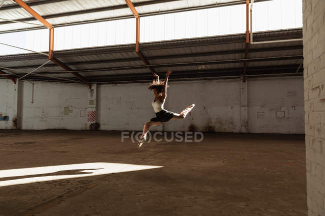 Side view of a young mixed race female ballet dancer wearing pointe shoes jumping in the air in shaft of sunlight with arms raised while dancing in an empty room at an abandoned warehouse — Stock Photo