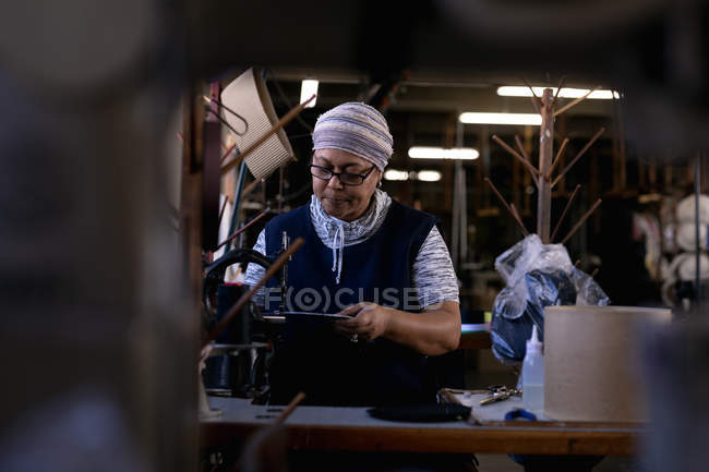 Front view of a middle aged mixed race woman using a sewing machine at a hat factory, surrounded by materials, seen through equipment — Stock Photo