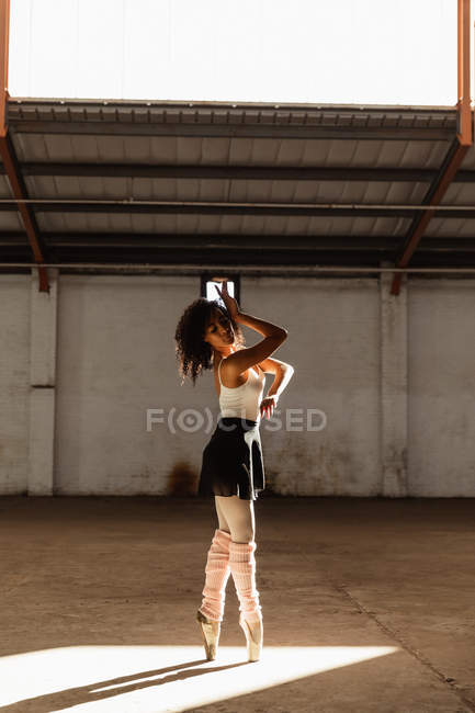 Seitenansicht einer jungen gemischten Rasse Balletttänzerin in Spitzenschuhen, die im Sonnenlicht auf ihren Zehen steht, während sie in einem leeren Raum einer verlassenen Lagerhalle tanzt — Stockfoto