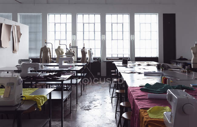 Vue de face des tables de travail avec des machines à coudre et des tissus dans un studio de design au collège de mode, avec des mannequins devant une fenêtre en arrière-plan — Photo de stock