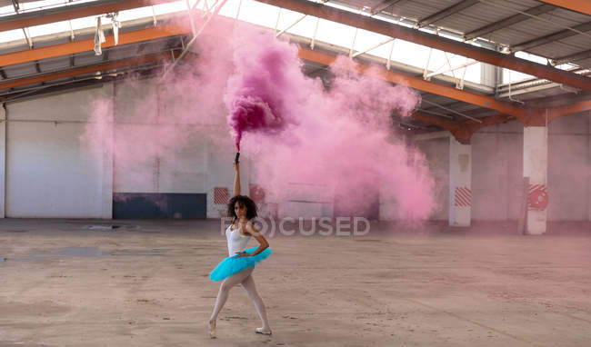 Vista frontal de una joven bailarina de ballet de raza mixta con un tutú azul y zapatos puntiagudos bailando sosteniendo una granada de humo rosa en una habitación vacía en un almacén abandonado - foto de stock