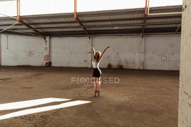 Вид збоку молодої змішаної раси танцівниці балету, яка стоїть на пальцях ніг з піднятими руками під час танців у порожній кімнаті на покинутому складі — стокове фото