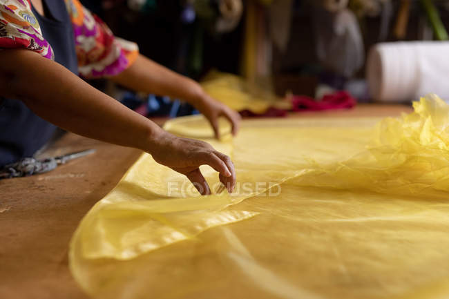 Vista laterale metà sezione di donna in piedi a un tavolo che lavora con tessuto giallo in una fabbrica di cappelli — Foto stock