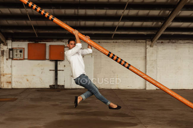 Side view of a young Caucasian male ballet dancer holding a structural pole and dancing with his feet off the ground in an empty room at an abandoned warehouse — Stock Photo