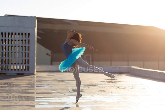 Vista laterale di una giovane ballerina di danza mista che indossa un tutù blu in piedi su una gamba sulle dita dei piedi in una posa da balletto, sul tetto di un edificio urbano, retroilluminato dalla luce del sole — Foto stock