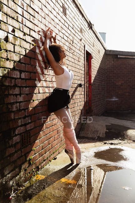Вид збоку крупним планом молода змішана раса танцівниця балету, що стоїть на пальцях на цегляній стіні з піднятими руками на даху міської будівлі — стокове фото