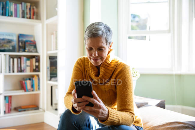 Vorderseite Nahaufnahme einer reifen kaukasischen Frau mit kurzen grauen Haaren, die zu Hause in ihrem Wohnzimmer sitzt und ein Smartphone benutzt, im Hintergrund ein sonnenbeschienenes Fenster — Stockfoto