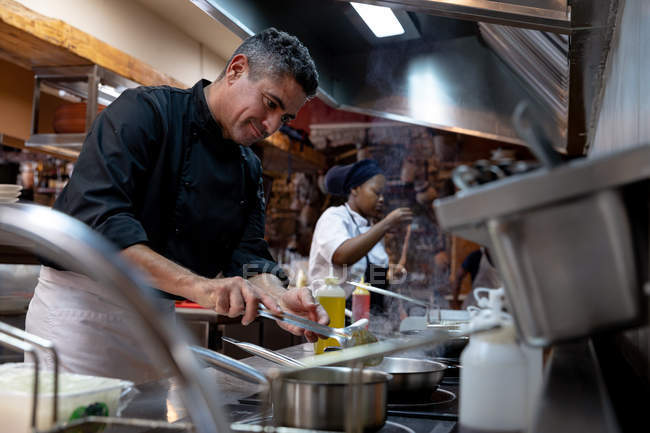Vista lateral de cerca de un chef caucásico de mediana edad cocinando comida en una cocina de restaurante ocupada, con otro personal de cocina trabajando en el fondo - foto de stock