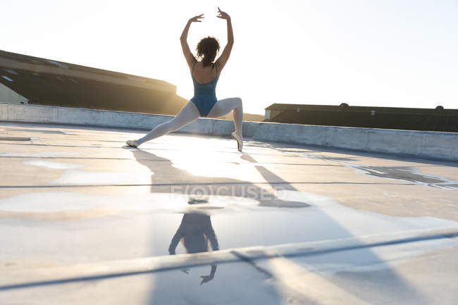 Visão traseira de uma jovem dançarina de balé mista em pé em uma pose de balé com os braços levantados, no telhado de um edifício urbano, retroiluminado pela luz solar e refletido na água da chuva — Fotografia de Stock
