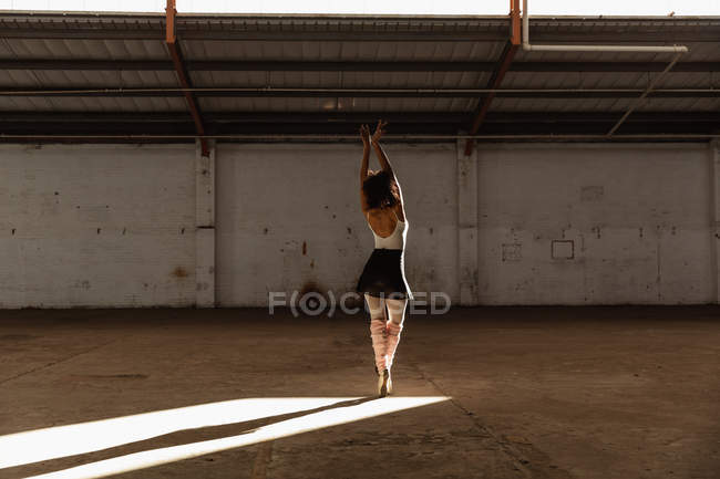Rückansicht einer jungen Balletttänzerin mit gemischter Rasse in Spitzenschuhen, die im Sonnenlicht auf den Zehenspitzen steht, mit erhobenen Armen, während sie in einem leeren Raum einer verlassenen Lagerhalle tanzt — Stockfoto