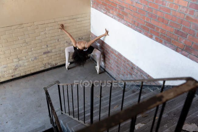Vista frontal elevada de una joven bailarina de ballet de raza mixta que sostiene una pose de baile en los dedos de los pies con los brazos levantados y la cabeza hacia abajo en una esquina en una escalera que aterriza en un almacén abandonado - foto de stock