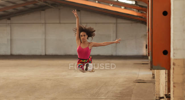 Vista frontal de una joven bailarina de ballet de raza mixta saltando en el aire con los brazos extendidos mientras baila en una habitación vacía en un almacén abandonado - foto de stock