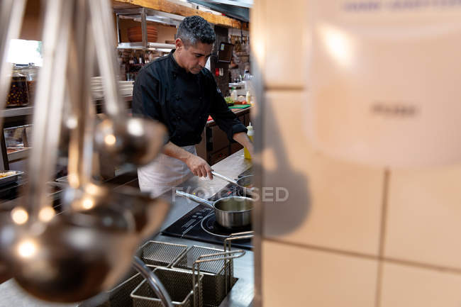Vista lateral de un chef caucásico de mediana edad cocinando con una sartén en una ajetreada cocina de restaurante, vista a través del equipo - foto de stock