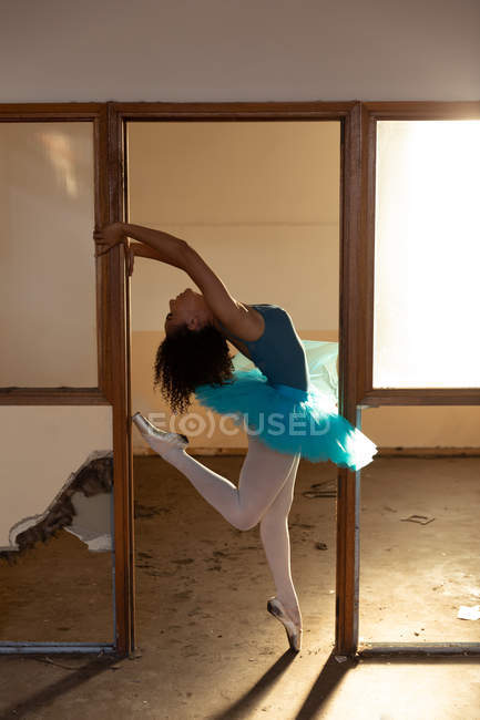 Вид сбоку молодой танцовщицы балета смешанной расы в синей пачке и пуантах, позирующей головой, стоящей на одной ноге на ногах в дверях заброшенного складского здания — стоковое фото