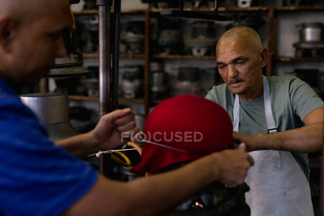 Side view of a senior and a middle aged mixed race men working together at a machine steaming the top of a hat to shape it in the workshop at a hat factory — Stock Photo
