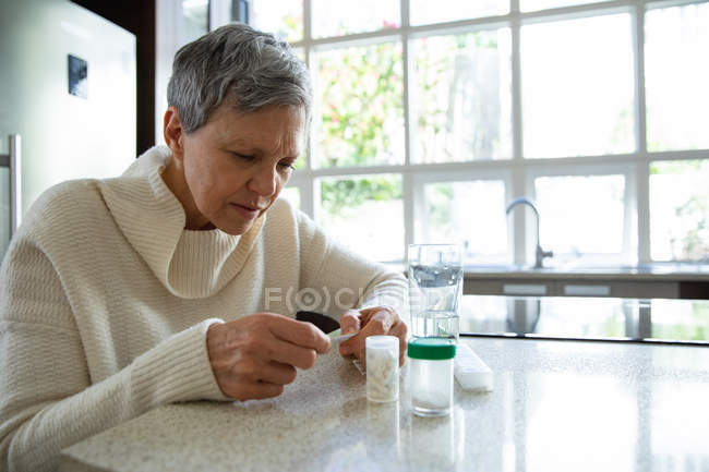 Seitenansicht einer reifen kaukasischen Frau mit kurzen grauen Haaren, die in ihrer Küche sitzt und ihre Medikamente betrachtet, mit Tablettenflaschen, einer wöchentlichen Tablettenbox und einem Glas Wasser auf dem Tresen neben ihr — Stockfoto