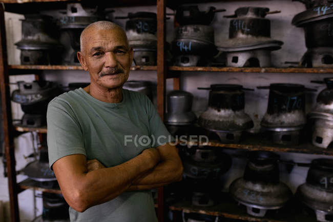 Retrato de un hombre de raza mixta sénior sonriente con los brazos cruzados, de pie frente a las estanterías de equipos tradicionales en el taller en una fábrica de sombreros - foto de stock