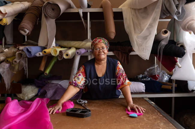 Портрет женщины среднего возраста смешанной расы, стоящей за столом, работающей с розовой тканью на шляпной фабрике и смотрящей в камеру улыбающейся . — стоковое фото