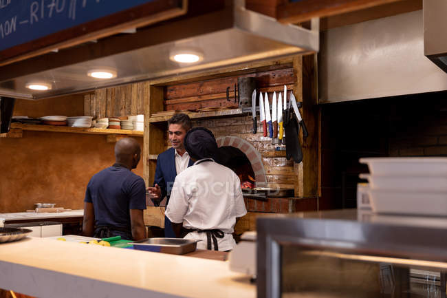 Front view of a middle aged Caucasian restaurant manager talking to two members of kitchen staff, seen from the back, in a restaurant kitchen — Stock Photo