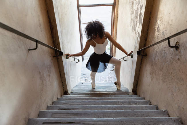 Erhöhte Frontansicht einer jungen Balletttänzerin mit gemischter Rasse, die das Geländer hält und unten auf einer Treppe in einer verlassenen Lagerhalle tanzt — Stockfoto