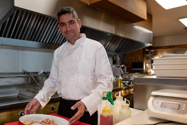 Porträt eines lächelnden kaukasischen Küchenchefs mittleren Alters, der in einer Restaurantküche einen Teller mit Essen hält — Stockfoto