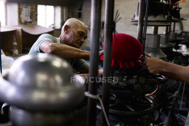 Seitenansicht eines Senior-Mixed-Race-Mannes, der in der Werkstatt einer Hutfabrik an einem Gerät arbeitet, das die Spitze eines Hutes formt, wobei Ausrüstung und die Hand eines Kollegen im Vordergrund sichtbar sind — Stockfoto