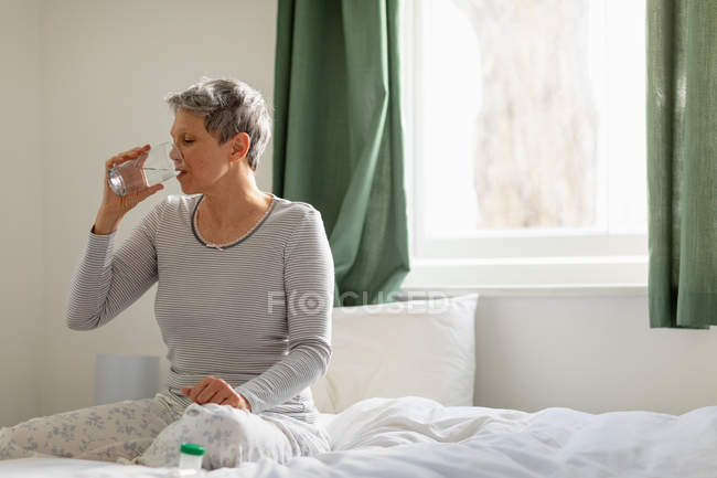 Vorderansicht einer reifen kaukasischen Frau mit kurzen grauen Haaren, die zu Hause auf ihrem Bett sitzt, ein Glas Wasser trinkt und Medikamente einnimmt, mit Behältern voller Medikamente auf dem Bett neben ihr — Stockfoto