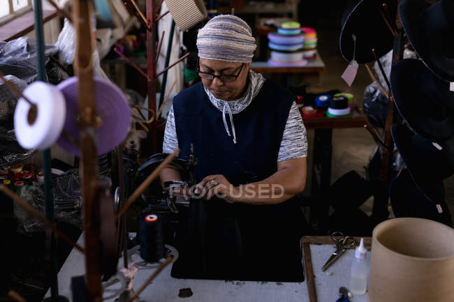 Vista frontale elevata di una donna di razza mista di mezza età che utilizza una macchina da cucire in una fabbrica di cappelli, circondata da materiali — Foto stock