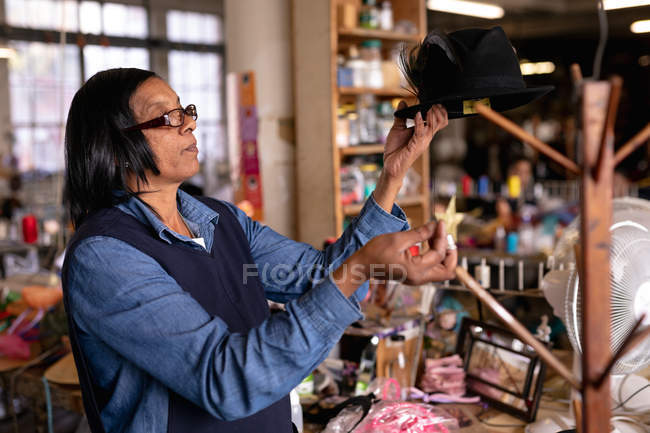 Vue latérale rapprochée d'une femme de race mixte d'âge moyen inspectant et ajoutant la touche finale à un chapeau sur un stand dans l'atelier d'une usine de chapeaux, avec des matériaux visibles en arrière-plan — Photo de stock