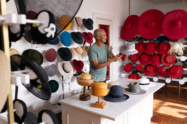 Vista lateral de un hombre de raza mixta senior de pie e inspeccionando un sombrero terminado, rodeado de sombreros en exhibición en la sala de exposición en una fábrica de sombreros - foto de stock