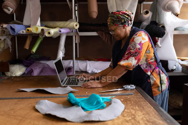Seitenansicht einer Frau mittleren Alters, die an einem Tisch steht und einen Laptop benutzt, während sie in einer Hutfabrik arbeitet. — Stockfoto