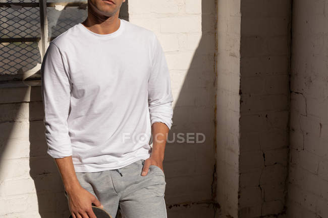 Vista frontal sección central de un joven parado contra una pared en un pozo de luz solar con una mano en el bolsillo, en un almacén abandonado - foto de stock
