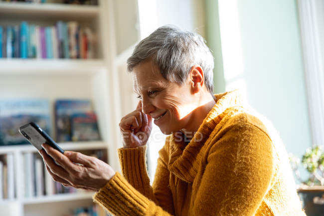 Vista lateral de cerca de una mujer caucásica madura con el pelo gris corto sentada en casa en su sala de estar sonriendo y usando un teléfono inteligente, iluminada por la luz del sol desde una ventana detrás de ella - foto de stock