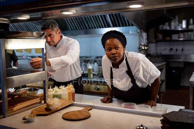 Front view close up of a middle aged Caucasian male chef checking orders at the order station while a young African American female chef stands waiting for food to be prepared for serving in a restaurant kitchen — Stock Photo