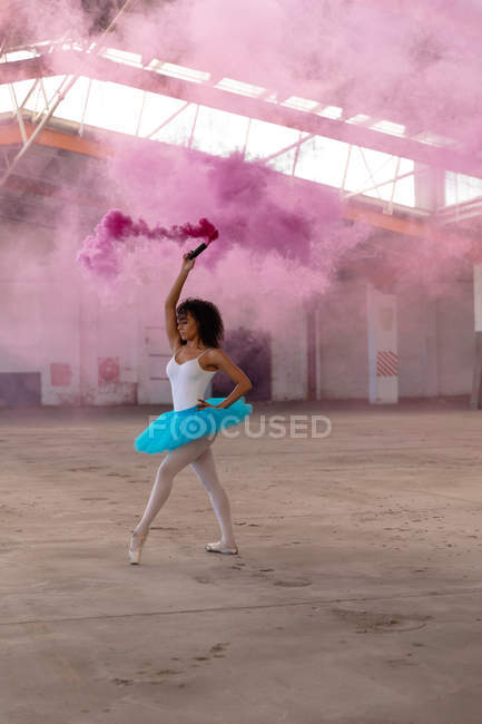 Вид збоку крупним планом молода змішана гонка жіноча танцівниця балету у блакитному туалеті та взутті, що танцює, тримаючи рожеву димову гранату у порожній кімнаті на покинутому складі — стокове фото