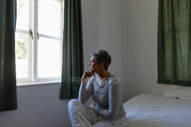Vista lateral de perto de uma mulher branca madura com cabelos brancos curtos sentados em sua cama e olhando pela janela em casa, com a mão no queixo — Fotografia de Stock