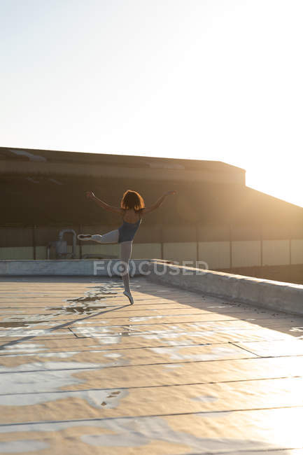 Вид сзади на молодую балетную танцовщицу смешанной расы, стоящую на одной ноге, с другой поднятой ногой и поднятыми руками, на крыше городского здания, освещенного солнечным светом — стоковое фото