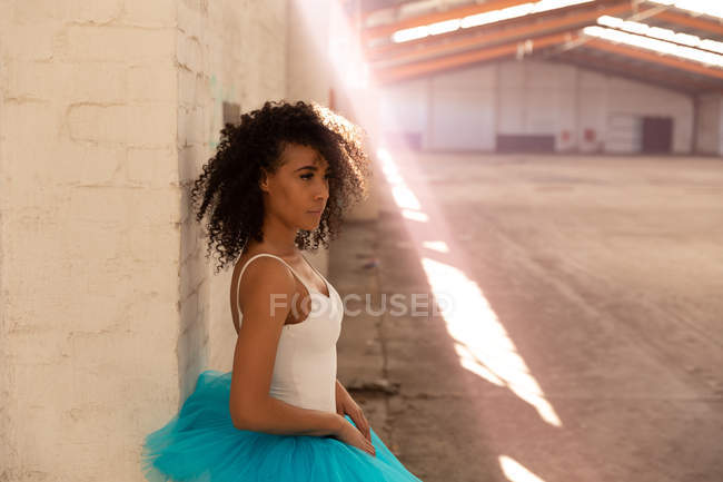 Seitenansicht einer jungen Balletttänzerin mit gemischter Rasse, die ein blaues Tutu trägt und vor einer Wand in einem leeren Raum einer verlassenen Lagerhalle steht, vor ihr ein Schacht mit Sonnenlicht — Stockfoto