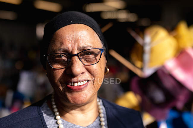 Портрет крупним планом жінка середнього віку змішаної раси посміхається до камери в майстерні на капелюшній фабриці, з барвистими капелюхами видно на задньому плані — стокове фото