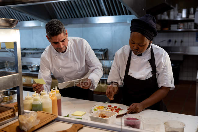 Front view close up of a middle aged Caucasian male chef checking orders while a young African American female chef stands prepares a dish for serving in a restaurant kitchen — Stock Photo