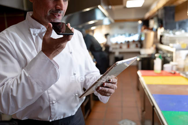 Вид спереди на белого повара средних лет, держащего в руках и разговаривающего по смартфону, пока он смотрит на планшетный компьютер, который держит в другой руке на кухне ресторана — стоковое фото