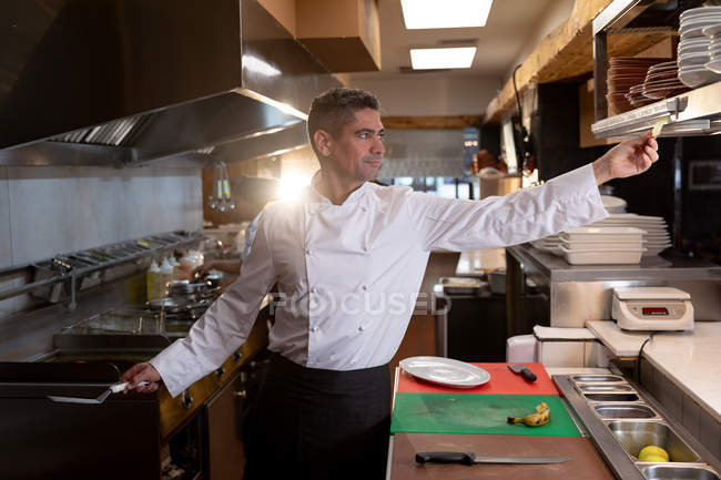 Front view close up of a middle aged Caucasian male chef reaching out to take an order at the order station in a restaurant kitchen — Stock Photo