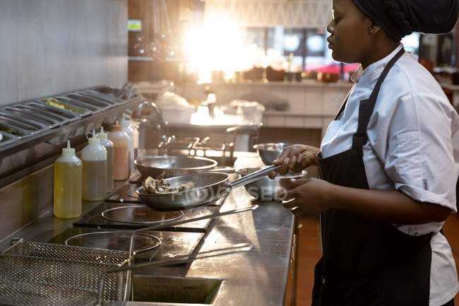 Vue de côté gros plan d'une jeune cuisinière afro-américaine qui fait frire des ingrédients dans une casserole dans une cuisine de restaurant — Photo de stock