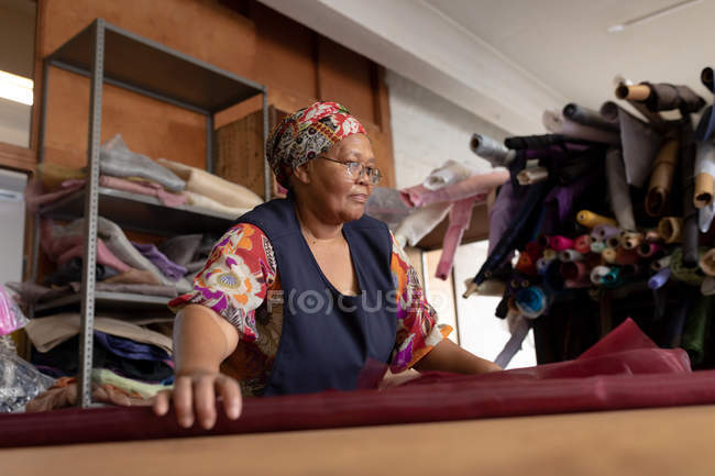 Vista frontale da vicino di una donna di razza mista di mezza età in piedi a un tavolo che lavora con tessuto in una fabbrica di cappelli . — Foto stock