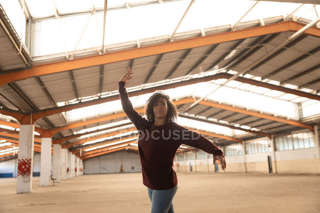 Vista frontale di una giovane ballerina di danza mista che indossa jeans e scarpe da punta che balla con le braccia tese in un magazzino abbandonato — Foto stock