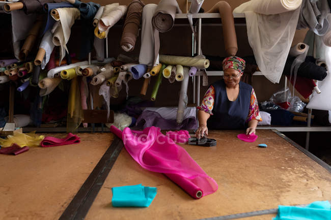 Vista frontale di una donna mista sorridente di mezza età in piedi a un tavolo che lavora con tessuto rosa in una fabbrica di cappelli . — Foto stock