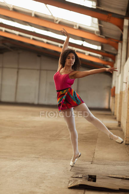 Vista frontal de cerca de una joven bailarina de ballet de raza mixta de pie sobre una pierna en los dedos de los pies con los brazos extendidos mientras baila en una habitación vacía en un almacén abandonado - foto de stock